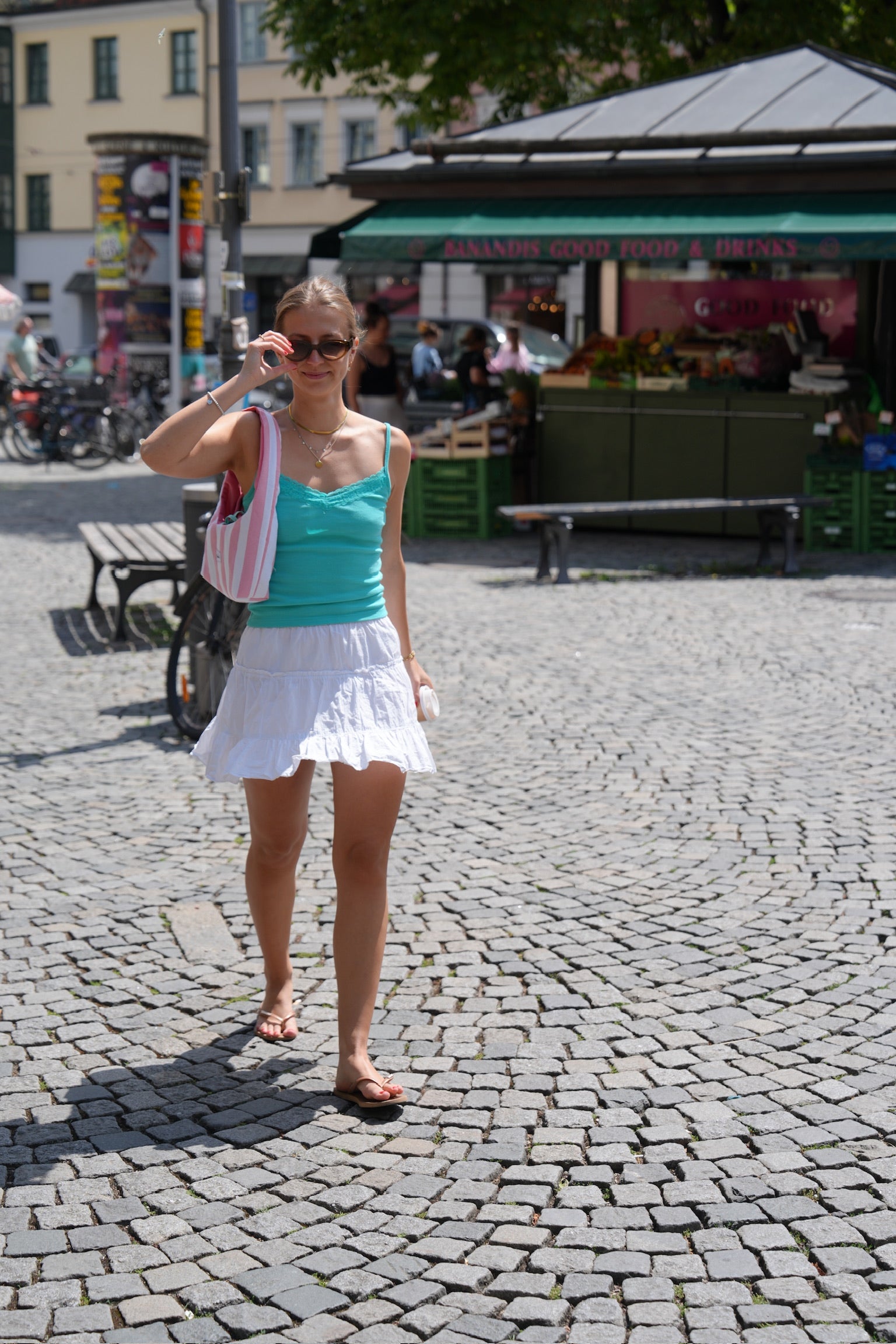 A woman walking on a cobblestone street wearing a turquoise tank top, a white ruffled mini skirt, and sunglasses, with a pink and white striped shoulder bag.