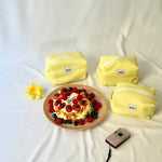 Three bright yellow cosmetic pouches with a soft, quilted knit-pattern fabric and “glow” logo labels arranged on a light fabric surface. In front of them is a small cake topped with strawberries, blueberries, and raspberries, alongside a yellow flower and a compact camera, creating a warm, summery and lifestyle-inspired scene.
