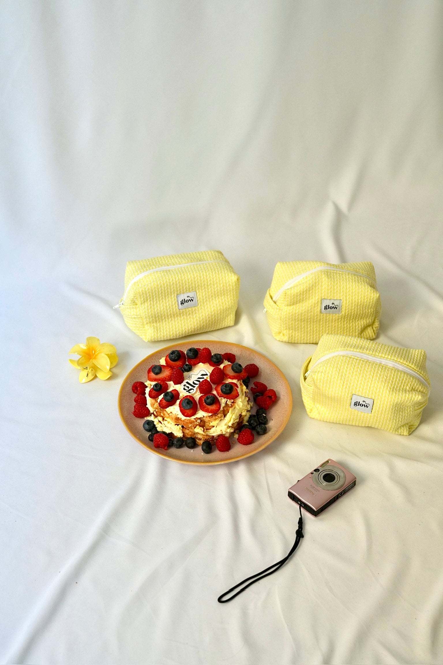 Three bright yellow cosmetic pouches with a soft, quilted knit-pattern fabric and “glow” logo labels arranged on a light fabric surface. In front of them is a small cake topped with strawberries, blueberries, and raspberries, alongside a yellow flower and a compact camera, creating a warm, summery and lifestyle-inspired scene.