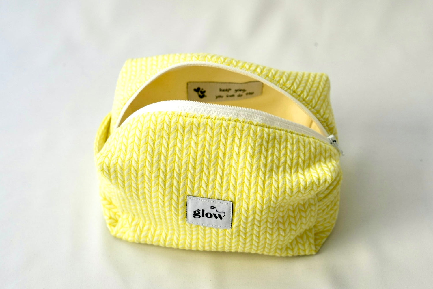 Open view of a bright yellow cosmetic pouch with a soft, quilted knit-pattern fabric and a white zipper. The bag reveals a spacious interior with a sewn-in label featuring a motivational message, combining functionality with an uplifting detail.