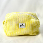 Front view of a soft, quilted cosmetic pouch in a bright yellow knit-pattern fabric with a white zipper. The bag features a centered “glow” logo label and a structured, padded shape, designed to hold everyday essentials.