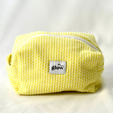 Front view of a soft, quilted cosmetic pouch in a bright yellow knit-pattern fabric with a white zipper. The bag features a centered “glow” logo label and a structured, padded shape, designed to hold everyday essentials.