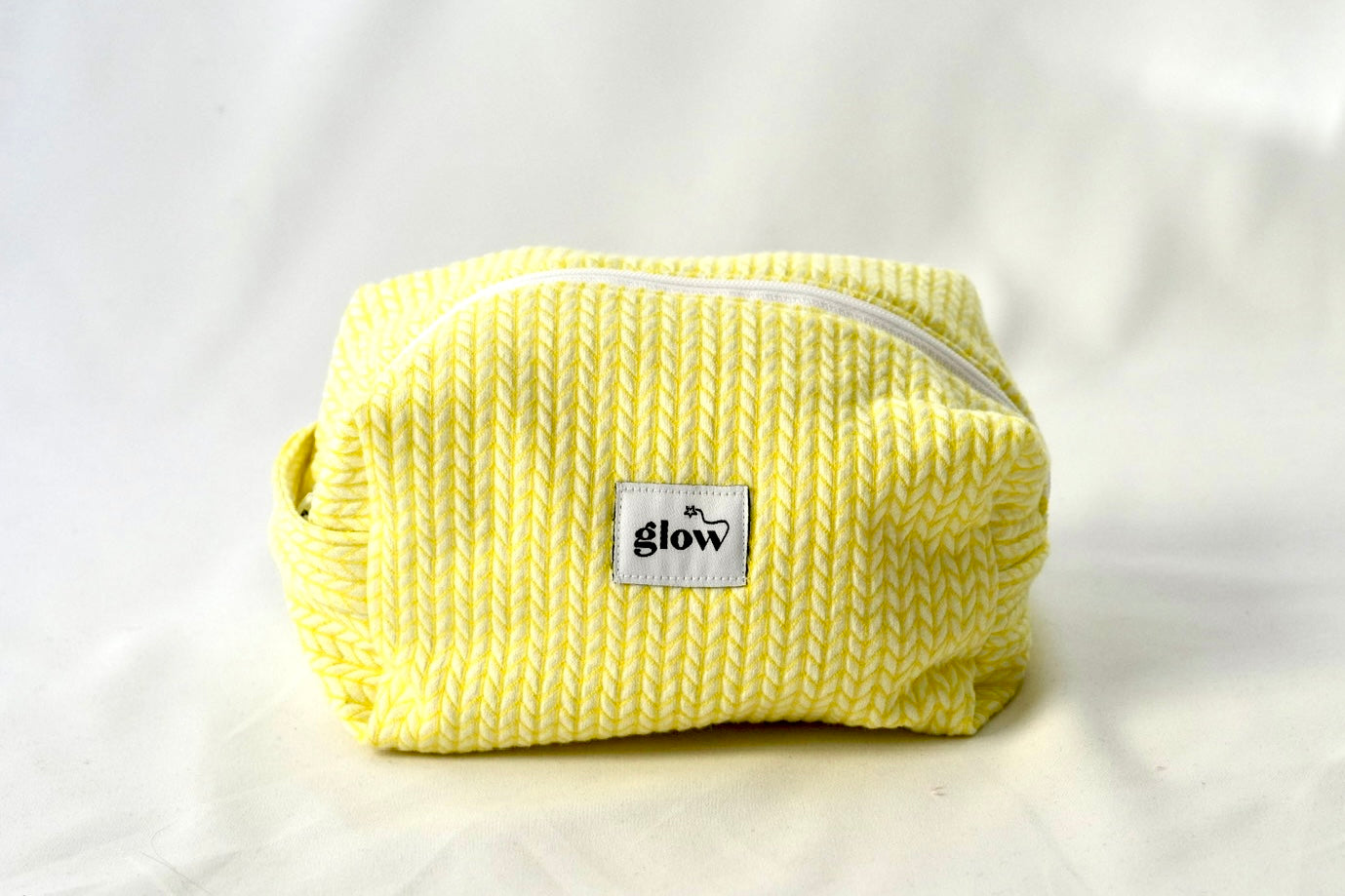Front view of a soft, quilted cosmetic pouch in a bright yellow knit-pattern fabric with a white zipper. The bag features a centered “glow” logo label and a structured, padded shape, designed to hold everyday essentials.