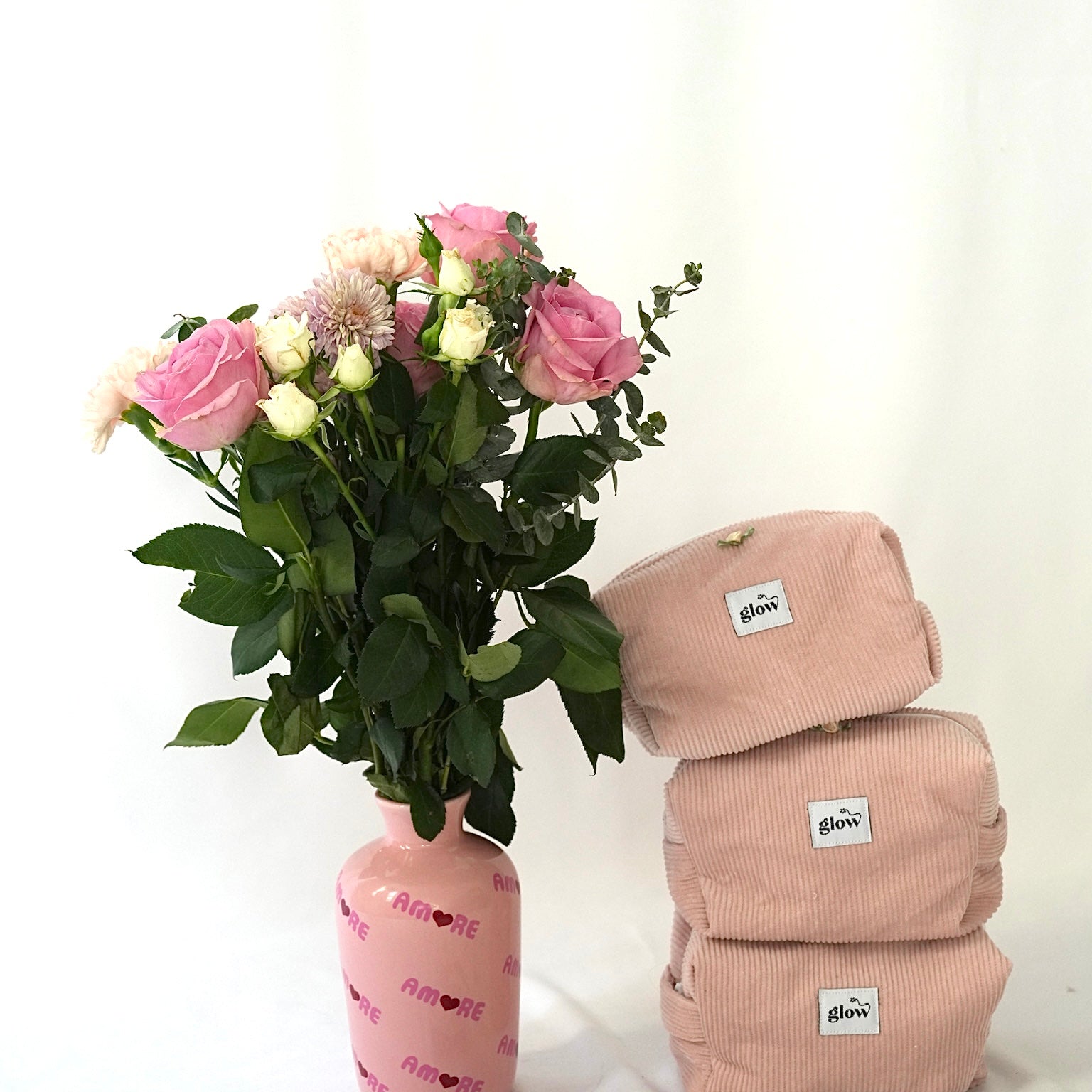 Three blush pink cosmetic pouches made from soft corduroy fabric with “glow” logo labels stacked next to a bouquet of pink and white flowers in a pink vase. A small camera and “glow” stickers are placed in front, creating a soft, feminine and lifestyle-inspired scene.