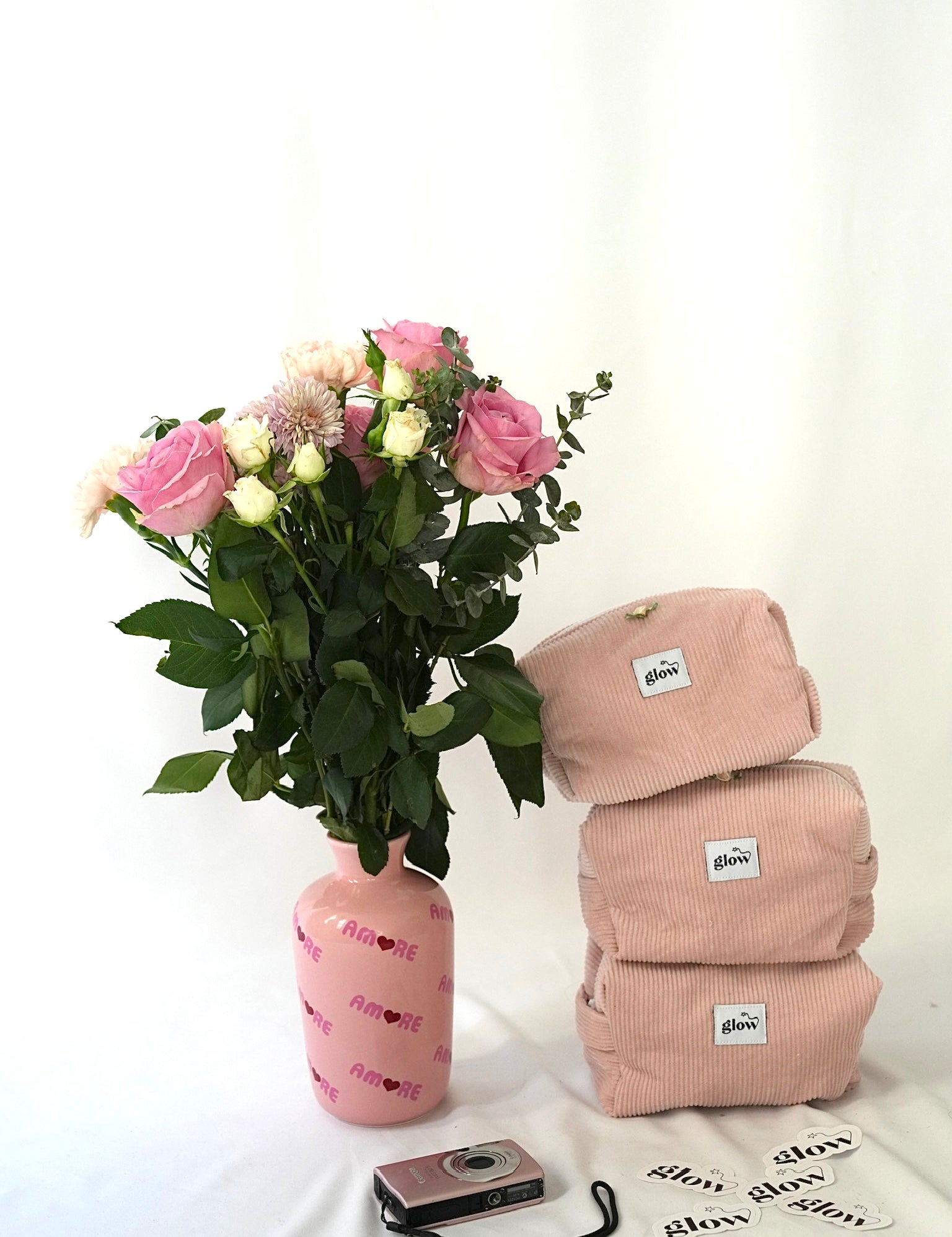 Three blush pink cosmetic pouches made from soft corduroy fabric with “glow” logo labels stacked next to a bouquet of pink and white flowers in a pink vase. A small camera and “glow” stickers are placed in front, creating a soft, feminine and lifestyle-inspired scene.