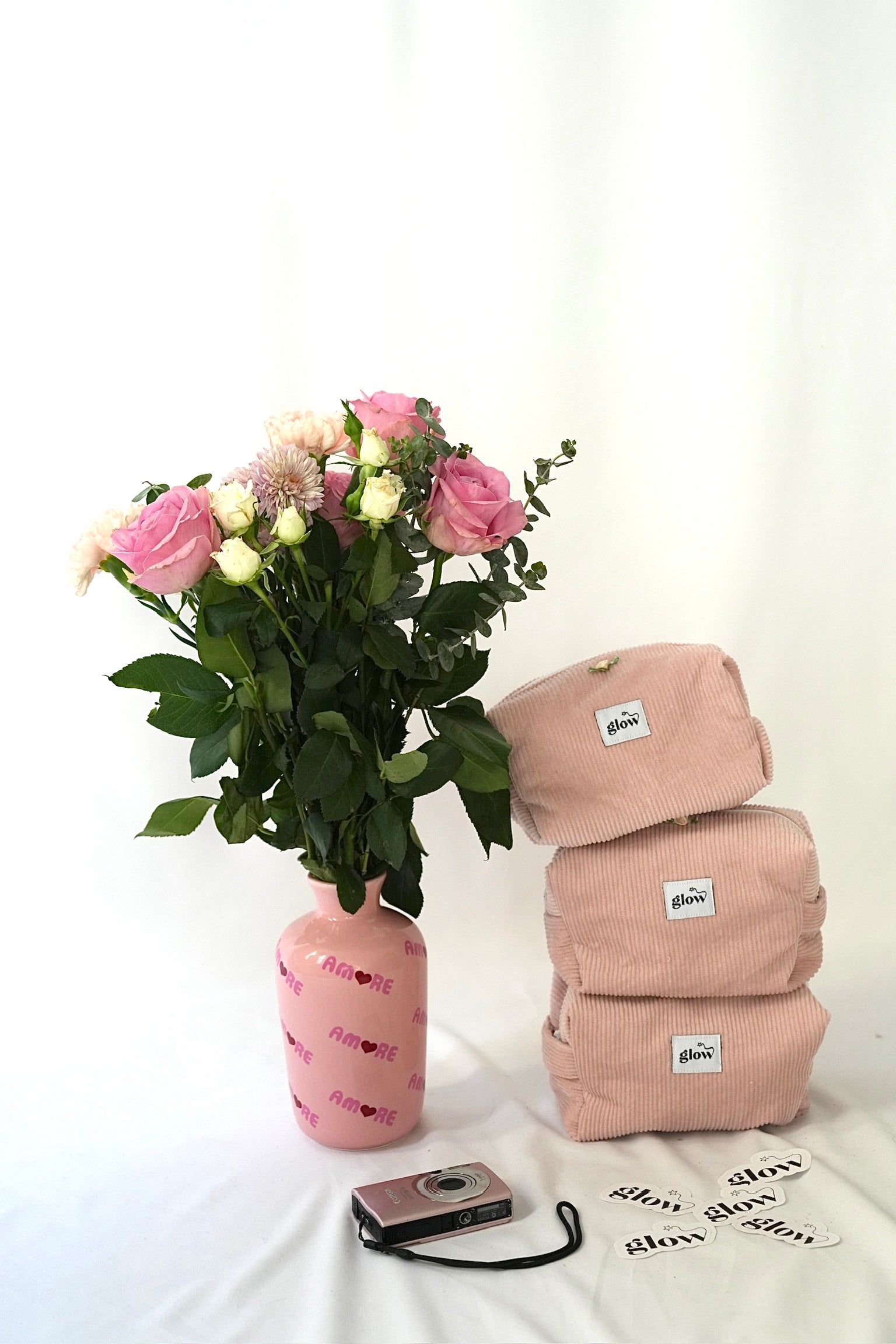 Three blush pink cosmetic pouches made from soft corduroy fabric with “glow” logo labels stacked next to a bouquet of pink and white flowers in a pink vase. A small camera and “glow” stickers are placed in front, creating a soft, feminine and lifestyle-inspired scene.