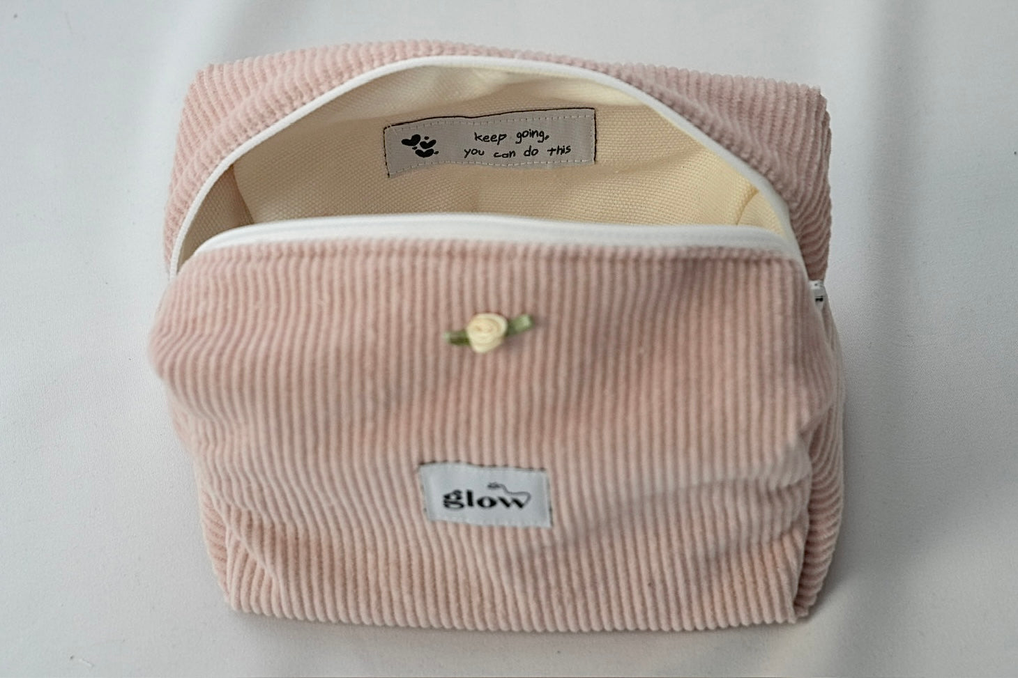 Open view of a blush pink cosmetic pouch made from soft corduroy fabric with a white zipper. The bag reveals a light-colored interior with a sewn-in label that reads “keep going, you can do this,” along with small heart details, combining functionality with an encouraging message.