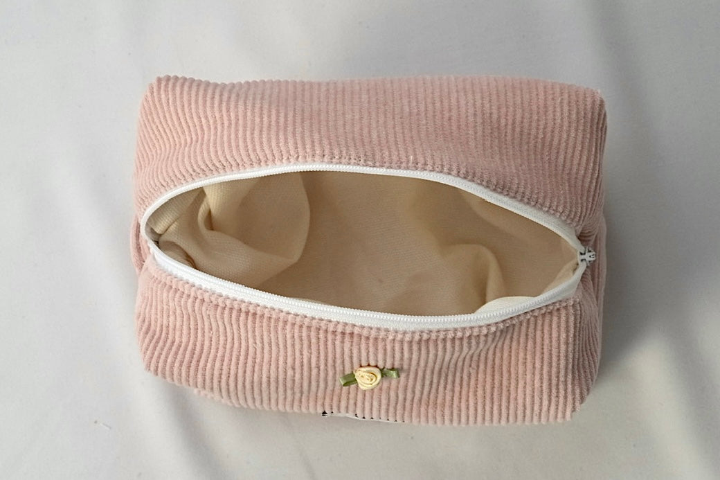 Open cosmetic pouch in blush pink corduroy fabric with a white zipper. The bag reveals a spacious, light-colored interior, highlighting its soft structure and practical design for everyday essentials.