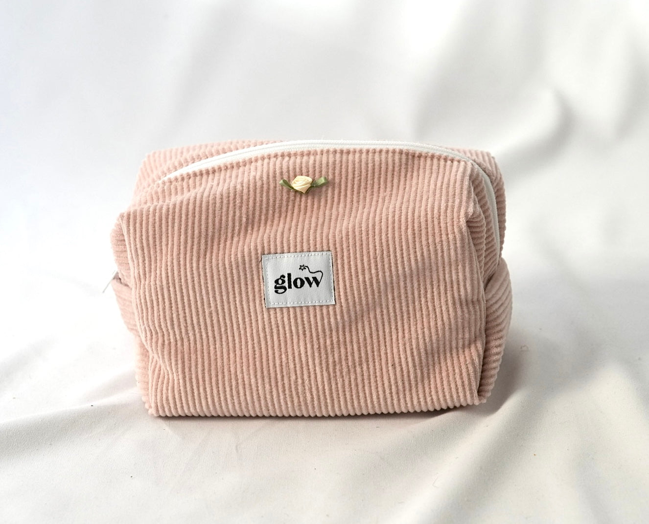 Front view of a soft, quilted cosmetic pouch in blush pink corduroy fabric with a white zipper. The bag features a centered “glow” logo label and a small decorative rose detail, creating a delicate and feminine design.