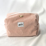 Front view of a soft, quilted cosmetic pouch in blush pink corduroy fabric with a white zipper. The bag features a centered “glow” logo label and a small decorative rose detail, creating a delicate and feminine design.