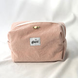 Front view of a soft, quilted cosmetic pouch in blush pink corduroy fabric with a white zipper. The bag features a centered “glow” logo label and a small decorative rose detail, creating a delicate and feminine design.