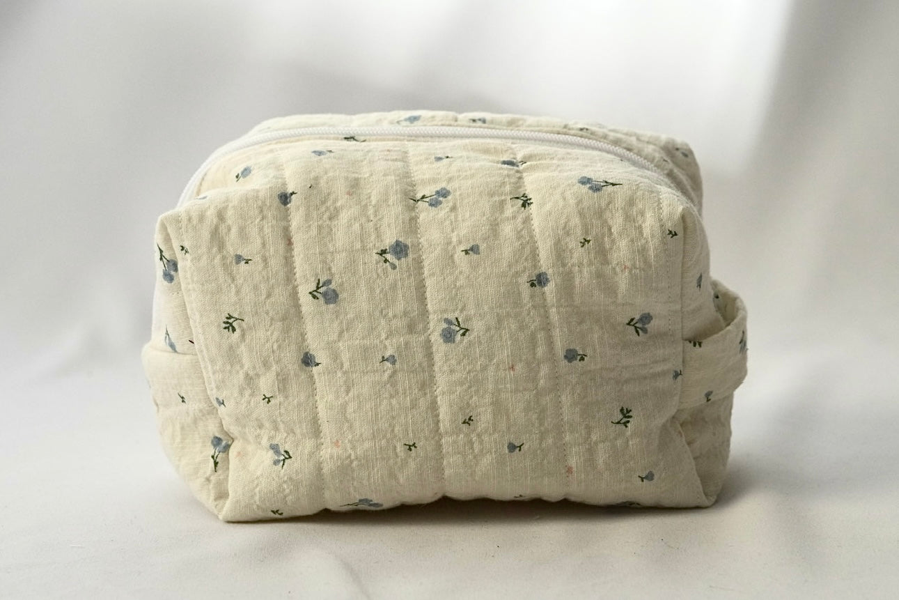 Back view of a soft, quilted cosmetic pouch in light cream fabric with a subtle blue floral pattern. The bag has a structured, padded shape with vertical stitching and a minimalist, label-free design.