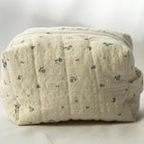 Back view of a soft, quilted cosmetic pouch in light cream fabric with a subtle blue floral pattern. The bag has a structured, padded shape with vertical stitching and a minimalist, label-free design.