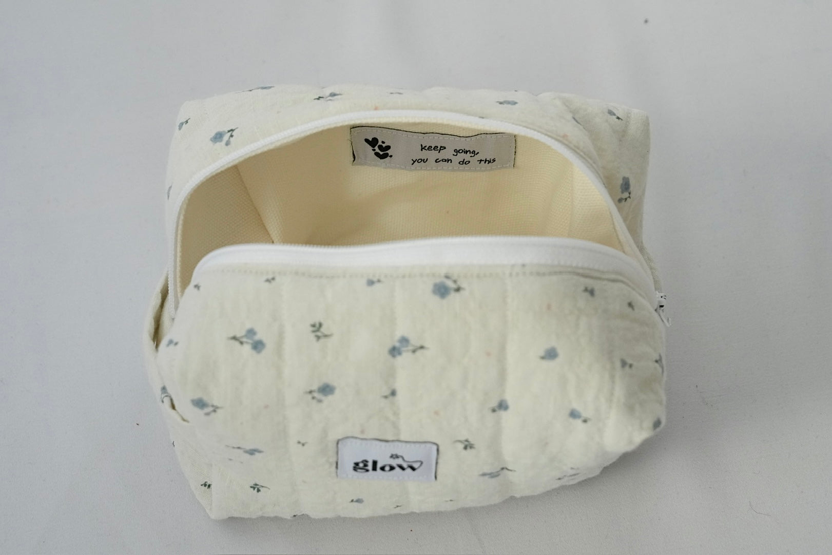 Open view of a soft, quilted cosmetic pouch in light cream fabric with a delicate blue floral pattern. The bag features a white zipper and a spacious interior with a sewn-in label that reads “keep going, you can do this,” alongside small heart icons.