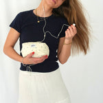 A woman holding a soft, quilted cosmetic pouch in light cream fabric with a delicate blue floral pattern and a “glow” logo label. She is wearing a navy t-shirt and white skirt, listening to wired earphones while gently tilting her head, creating a relaxed and lifestyle-focused scene.