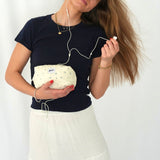 A woman holding a soft, quilted cosmetic pouch in light cream fabric with a delicate blue floral pattern and a “glow” logo label. She is wearing a navy t-shirt and white skirt, listening to wired earphones while gently tilting her head, creating a relaxed and lifestyle-focused scene.