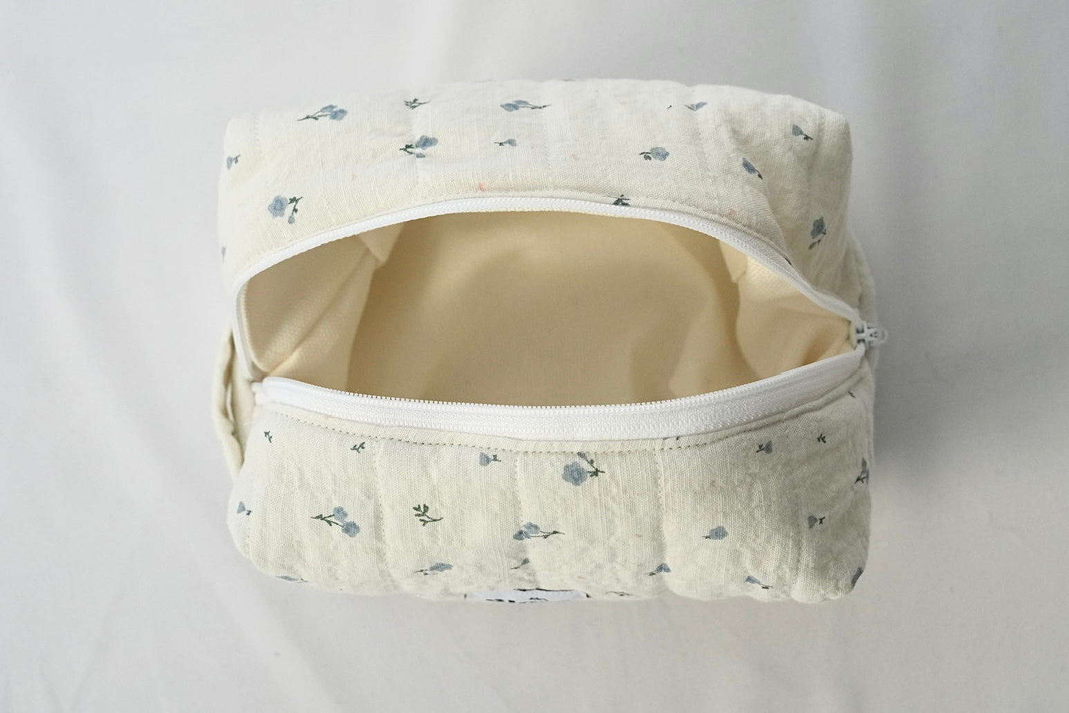Open cosmetic pouch made of soft, quilted cream fabric with a delicate blue floral pattern and a white zipper. The bag reveals a spacious, light-colored interior designed to hold everyday essentials.