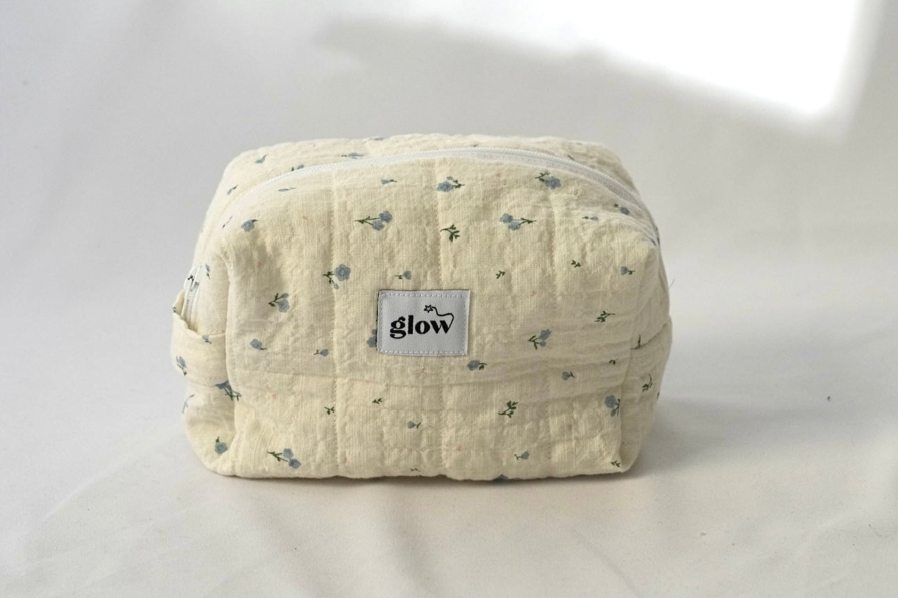 A soft, quilted cosmetic pouch in light cream fabric with a delicate blue floral pattern and a white zipper. The front features a small “glow” logo label. The bag has a structured, spacious shape, perfect for storing everyday essentials.
