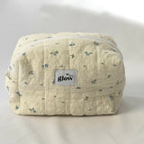 A soft, quilted cosmetic pouch in light cream fabric with a delicate blue floral pattern and a white zipper. The front features a small “glow” logo label. The bag has a structured, spacious shape, perfect for storing everyday essentials.