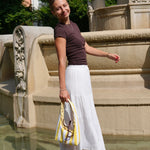 A woman stands on a stone ledge in front of a fountain, wearing a dark brown short-sleeve top, a long white tiered skirt, and holding a yellow and white striped tote bag.