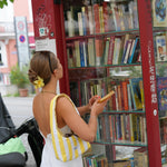 A woman in a white dress and yellow striped shoulder bag stands in front of a red glass bookshelf filled with books, holding a book in a street setting.