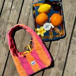 A striped terrycloth shoulder bag in pink, orange, and yellow lies on a weathered wooden deck next to a blue tray holding lemons, oranges, sunglasses, and a small flower.