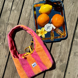 A striped terrycloth shoulder bag in pink, orange, and yellow lies on a weathered wooden deck next to a blue tray holding lemons, oranges, sunglasses, and a small flower.