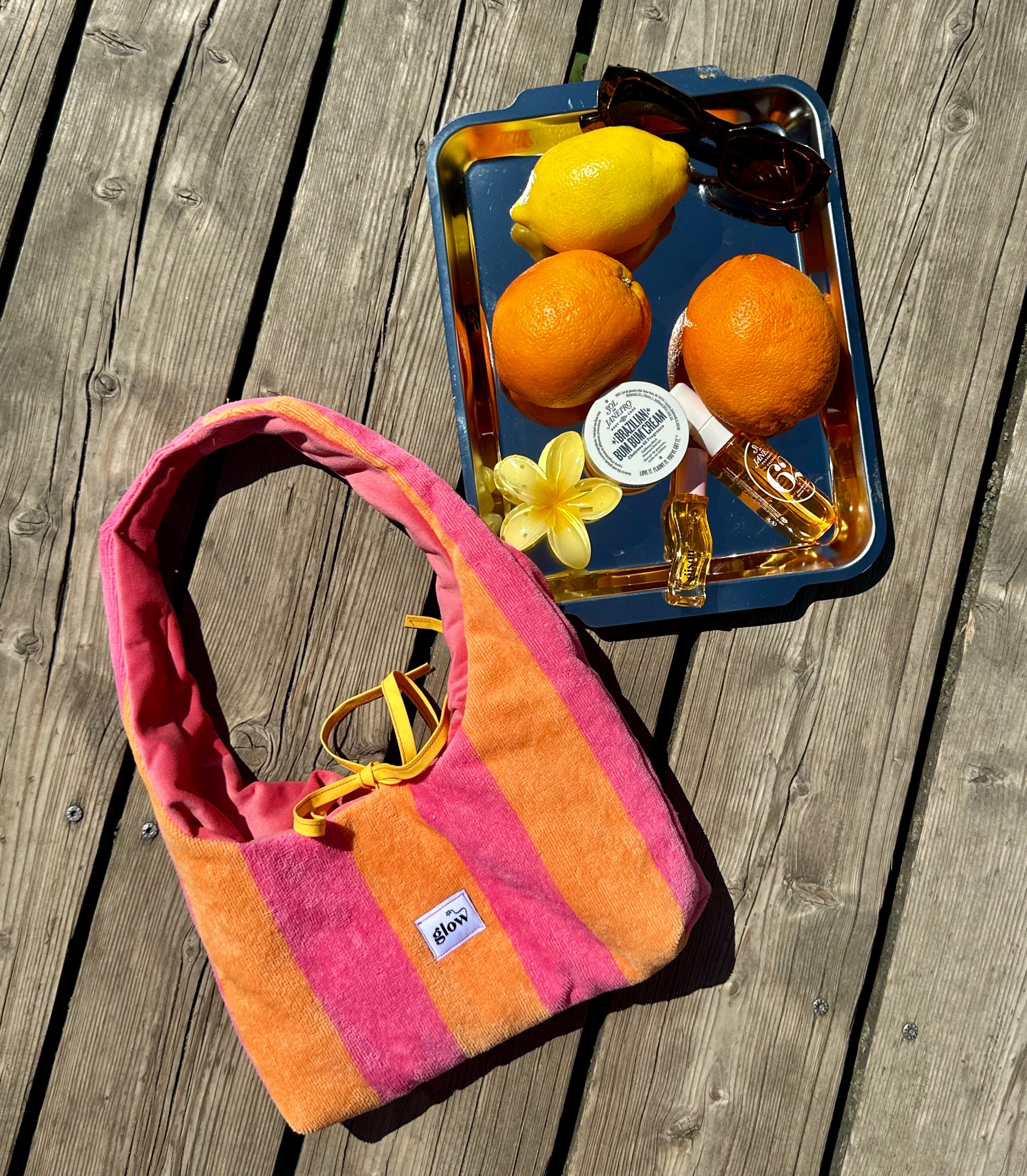 A striped terrycloth shoulder bag in pink, orange, and yellow lies on a weathered wooden deck next to a blue tray holding lemons, oranges, sunglasses, and a small flower.