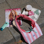 A pink and white striped fabric shoulder bag with a mint green ribbon tie, styled on gray stone pavement next to a Bazaar magazine, a tube of lip balm, a coffee cup, and a small tray of accessories.