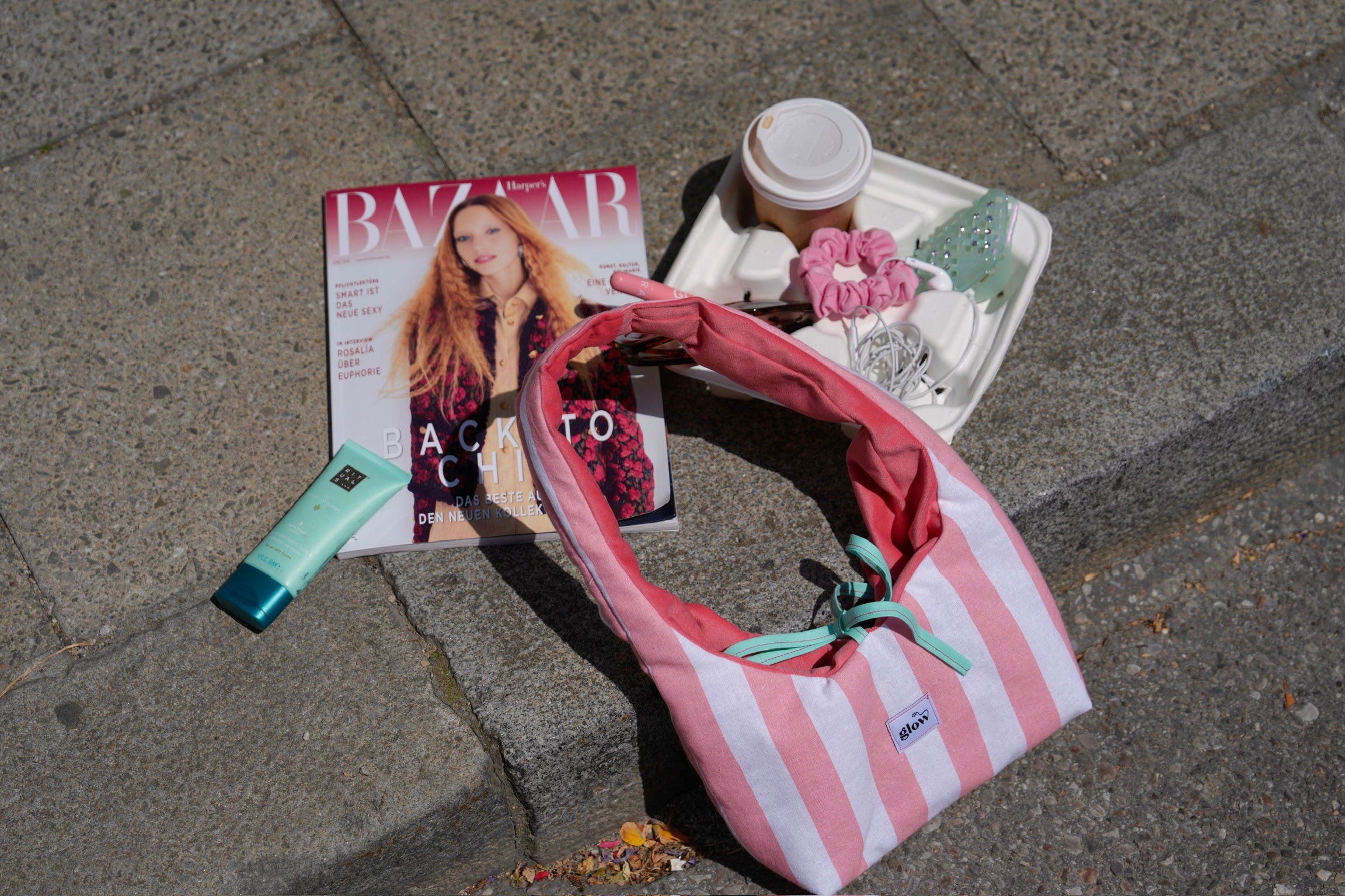 A pink and white striped fabric shoulder bag with a mint green ribbon tie, styled on gray stone pavement next to a Bazaar magazine, a tube of lip balm, a coffee cup, and a small tray of accessories.