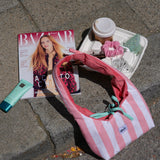 A pink and white striped fabric shoulder bag with a mint green ribbon tie, styled on gray stone pavement next to a Bazaar magazine, a tube of lip balm, a coffee cup, and a small tray of accessories.