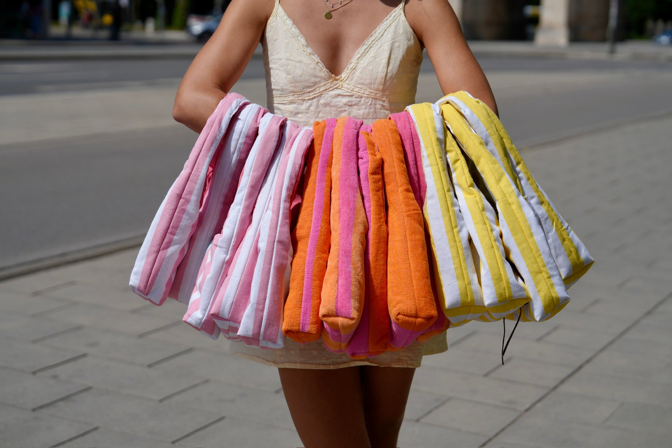 A person outdoors holding three sets of striped bath towels in pink, orange, and yellow, wearing a beige dress.