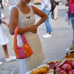 A woman in a white sleeveless dress stands at an outdoor fruit market stall, holding a pink and orange shoulder bag. Fresh fruits including dragon fruit, bananas, and melons are displayed in wooden bowls on the counter, with other shoppers blurred in the background.