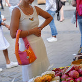 A woman in a white sleeveless dress stands at an outdoor fruit market stall, holding a pink and orange shoulder bag. Fresh fruits including dragon fruit, bananas, and melons are displayed in wooden bowls on the counter, with other shoppers blurred in the background.