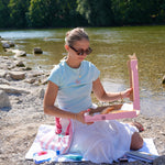 A woman sitting on a blanket on a rocky riverbank, holding a pink pizza box with pizza inside. She is wearing a light blue t-shirt and a white skirt, with trees and water in the background.