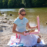 A woman sitting on a blanket on a rocky riverbank, holding a pink pizza box with pizza inside. She is wearing a light blue t-shirt and a white skirt, with trees and water in the background.