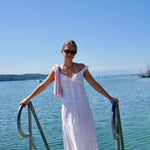 A woman wearing a white tiered maxi dress and sunglasses stands barefoot on a wooden dock railing overlooking a calm blue-green body of water under a clear blue sky.