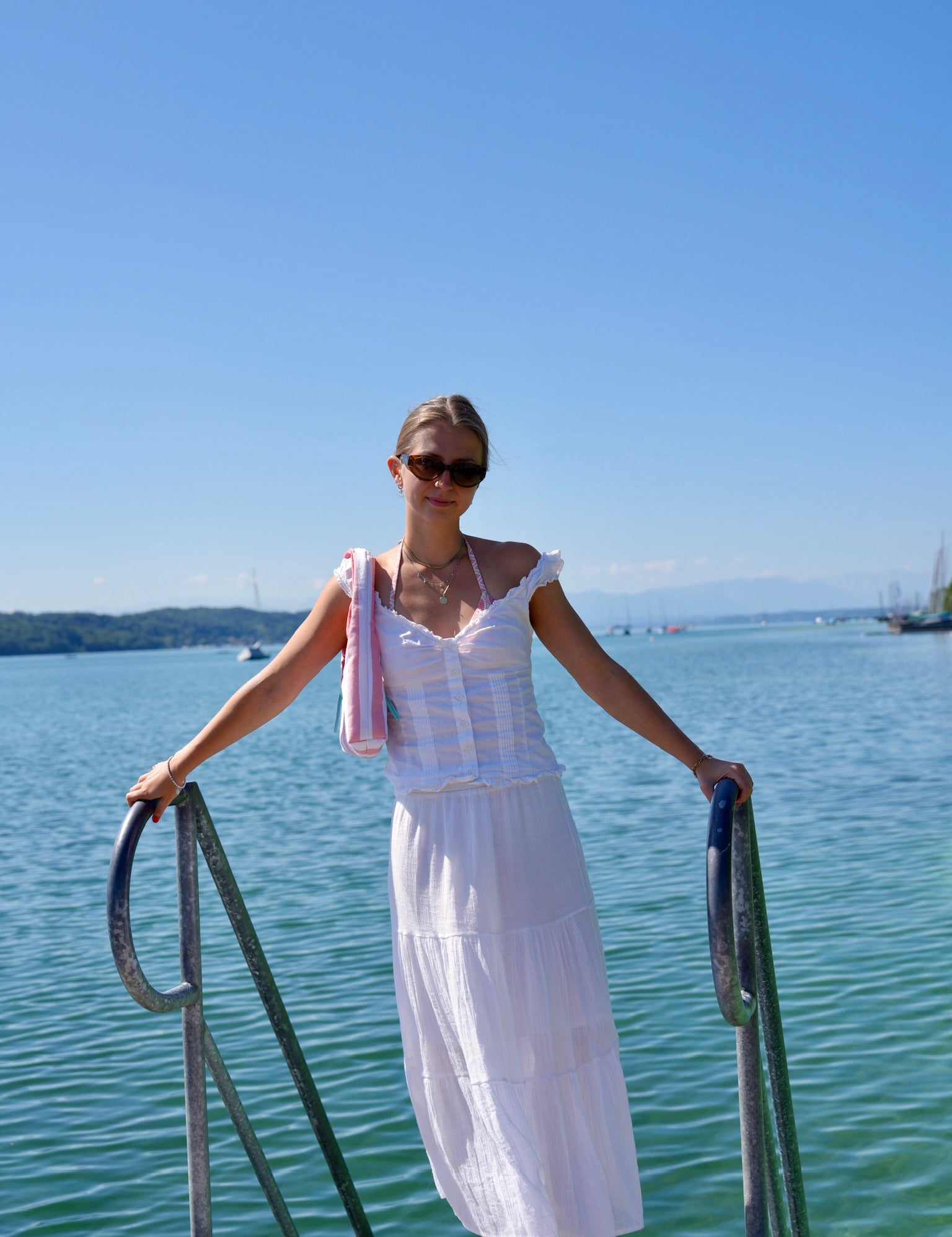 A woman wearing a white tiered maxi dress and sunglasses stands barefoot on a wooden dock railing overlooking a calm blue-green body of water under a clear blue sky.