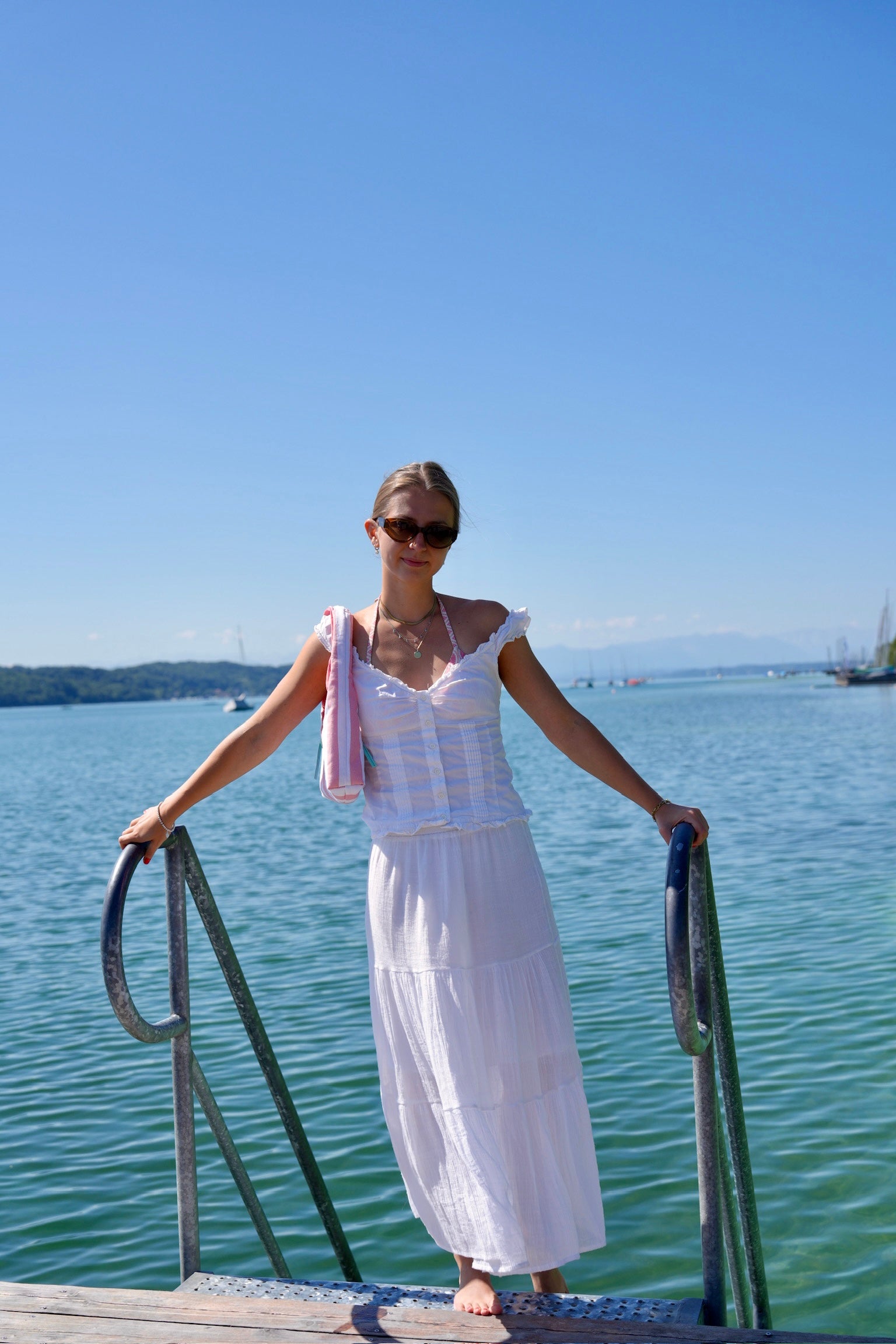 A woman wearing a white tiered maxi dress and sunglasses stands barefoot on a wooden dock railing overlooking a calm blue-green body of water under a clear blue sky.