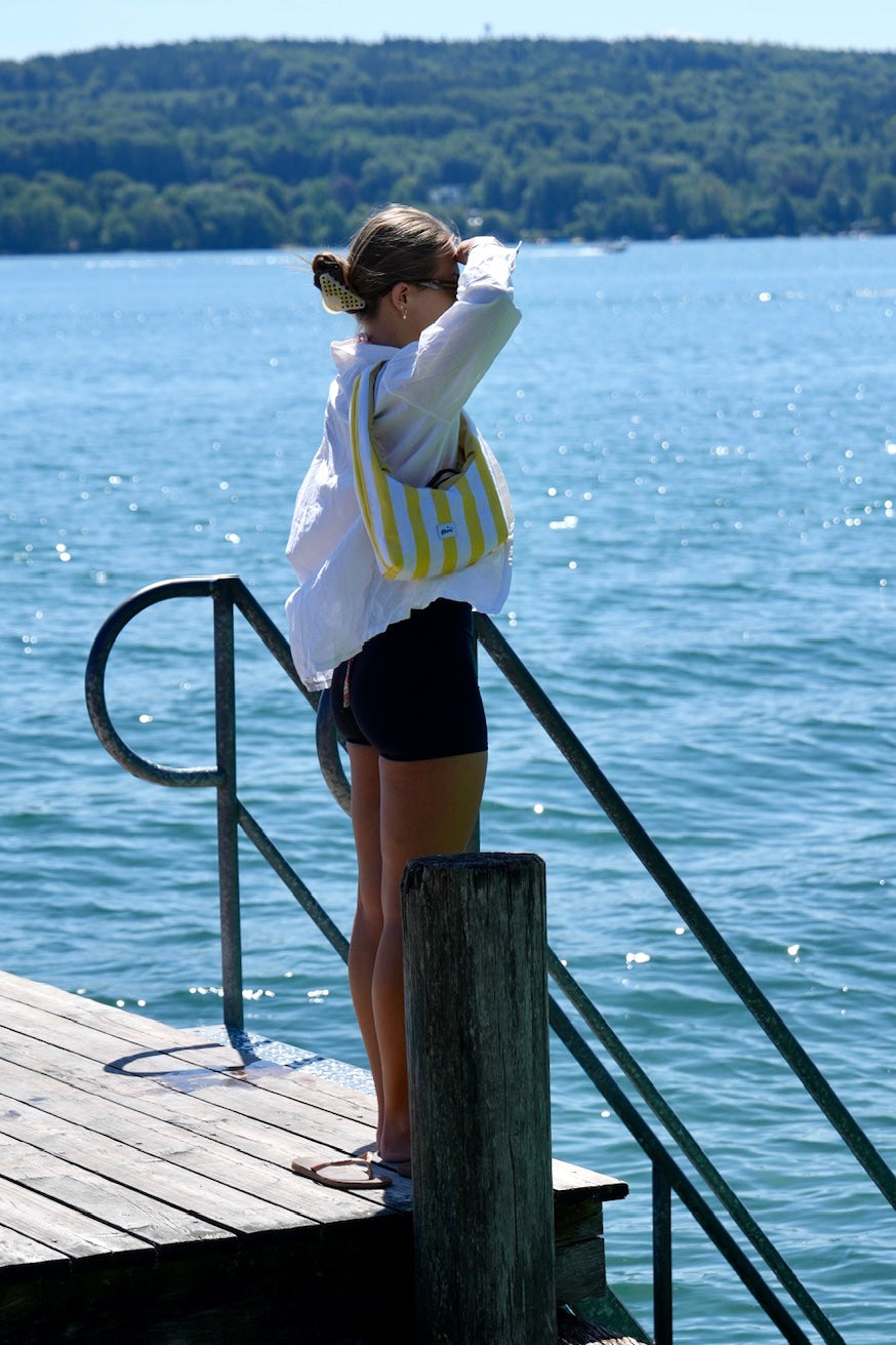 A woman stands on a wooden dock by a large body of water, wearing a white long-sleeve shirt, black shorts, and carrying a yellow and white striped shoulder bag.