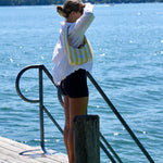 A woman stands on a wooden dock by a large body of water, wearing a white long-sleeve shirt, black shorts, and carrying a yellow and white striped shoulder bag.
