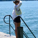 A woman stands on a wooden dock by a large body of water, wearing a white long-sleeve shirt, black shorts, and carrying a yellow and white striped shoulder bag.
