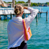 A woman seen from behind wearing a white long-sleeve shirt and black shorts, carrying a bright pink and orange shoulder bag while standing on a wooden dock over turquoise water.