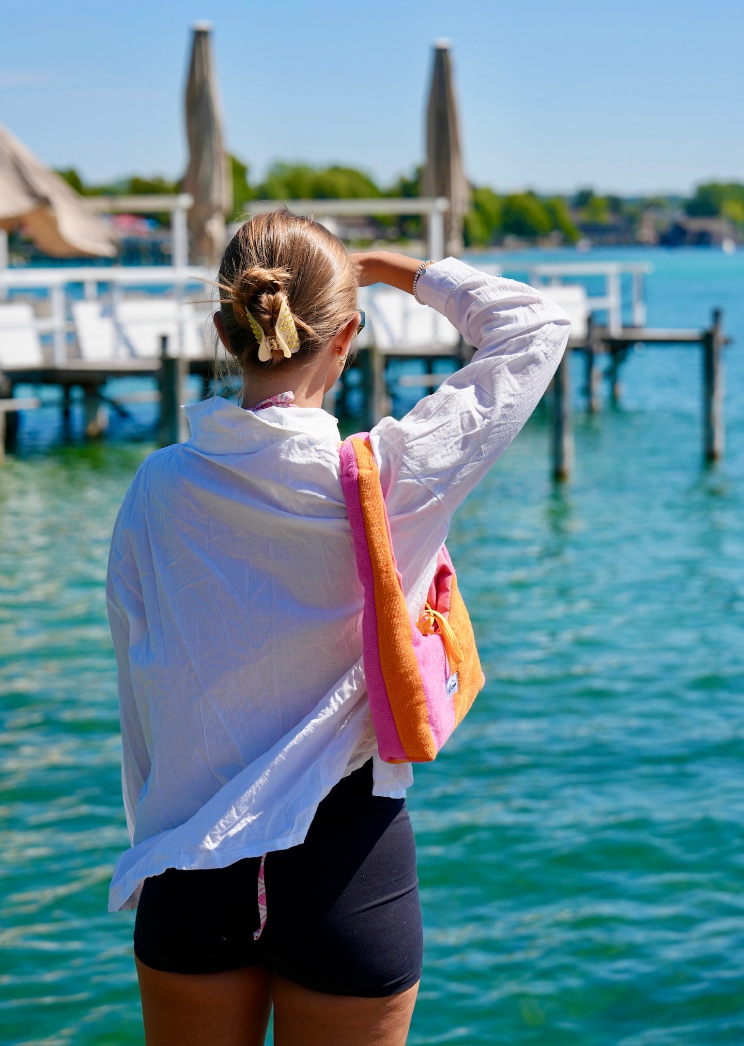 A woman seen from behind wearing a white long-sleeve shirt and black shorts, carrying a bright pink and orange shoulder bag while standing on a wooden dock over turquoise water.