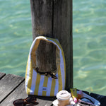 A yellow and white striped shoulder bag with a small blue logo, resting on a weathered wooden dock next to a wooden post with clear turquoise water in the background. A small snack tray, sunglasses, and a coffee cup are also on the dock.