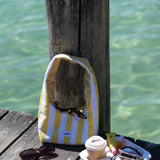 A yellow and white striped shoulder bag with a small blue logo, resting on a weathered wooden dock next to a wooden post with clear turquoise water in the background. A small snack tray, sunglasses, and a coffee cup are also on the dock.