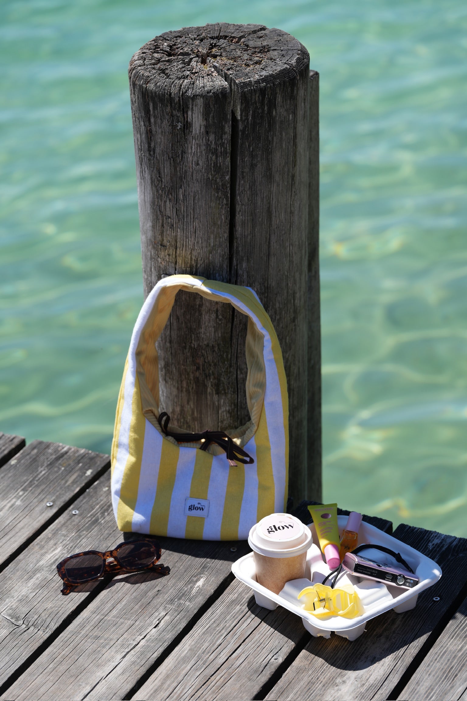 A yellow and white striped shoulder bag with a small blue logo, resting on a weathered wooden dock next to a wooden post with clear turquoise water in the background. A small snack tray, sunglasses, and a coffee cup are also on the dock.