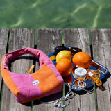 A pink and orange striped fabric shoulder bag with a white 'glow' label, styled on a weathered wooden dock with oranges, sunglasses, earphones, and a small jar against a background of green water.