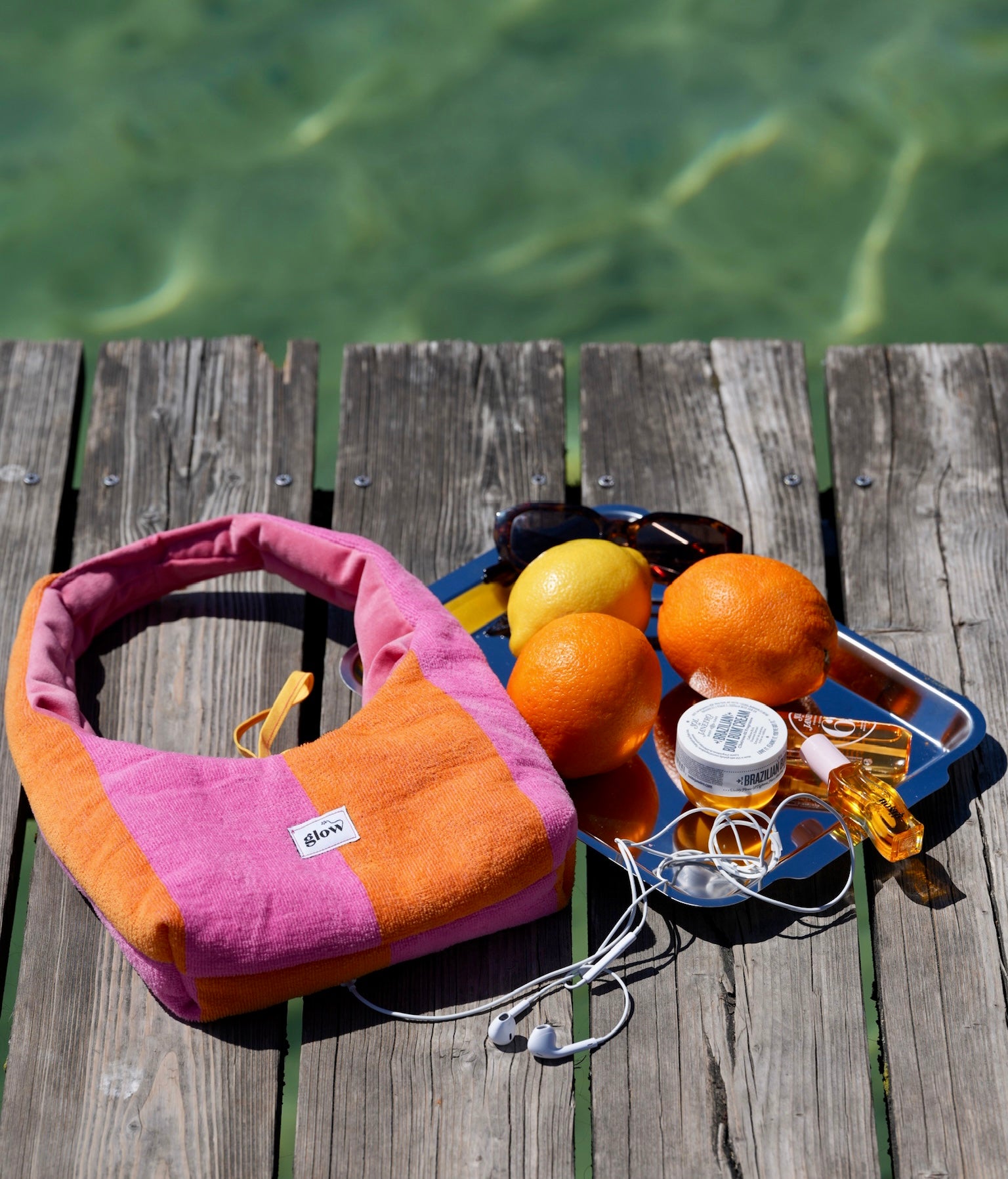 A pink and orange striped fabric shoulder bag with a white 'glow' label, styled on a weathered wooden dock with oranges, sunglasses, earphones, and a small jar against a background of green water.
