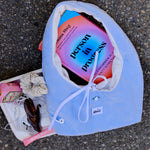 A light blue fabric pouch with a white drawstring closure, laid on asphalt next to a white tray holding sunglasses, a scrunchie, and a book titled 'Person in progress'. The pouch has a small white tag reading 'ilora'.
